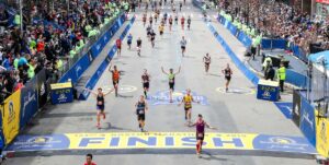 A photograph taken slightly above the finish line of the Boston Marathon. The runners are spaced out as they cross the finish line and are rejoicing with their hands in the air. You can see the large crowds of spectators cheering on the sidelines. 
